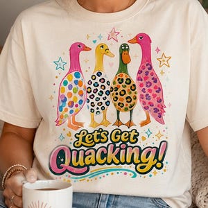 Peut inclure: Un t-shirt à manches courtes de couleur crème avec un motif représentant quatre canards aux corps colorés et à motifs. Le texte "Let's Get Quacking!" est imprimé sous les canards dans une police audacieuse et multicolore, entouré d'étoiles.