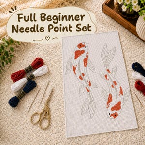 May include: A needlepoint kit featuring a rectangular canvas with two koi fish designs in orange and white. The set includes three skeins of embroidery floss in red, white, and navy blue, two metal needles, and gold-toned decorative scissors.