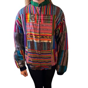 May include: A long-sleeve zip-up jacket featuring a patchwork design of multicolored striped fabric panels. The garment includes a green stand-up collar, a central metal zipper closure, and dark blue ribbed cuffs at the wrists and waist hem.