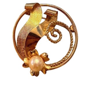 May include: A circular gold-tone brooch featuring a central flower motif with a single round cream-colored pearl. The design includes a textured ribbon-like element and a twisted rope-style wire accent, all enclosed within a smooth circular frame.