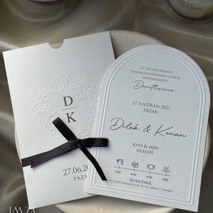 May include: Elegant white wedding invitation set with embossed floral details and a black satin ribbon. The invitation features the initials "D K" and event details. The set includes a card with an arched top and event information.