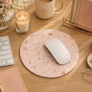 May include: A circular mouse pad featuring a light pink terrazzo pattern with scattered gold flecks. The surface has a smooth, speckled appearance with various shades of pink and white fragments embedded throughout the design, providing a flat base.