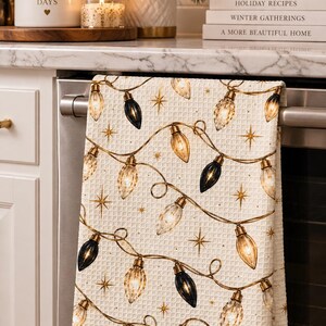 May include: A cream-colored waffle-weave kitchen towel featuring a repeating pattern of hanging string lights with gold and black bulbs, interspersed with small gold starburst accents, draped over the handle of a stainless steel oven.