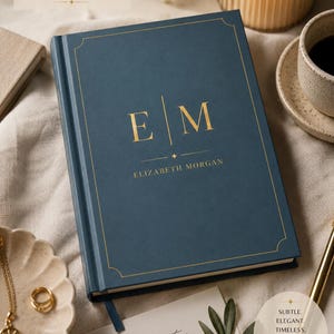 May include: A dark blue hardback journal featuring gold foil lettering with the initials E M and the name ELIZABETH MORGAN centred on the front cover. The cover includes a thin gold border and a small star icon above the name.