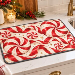 Puede incluir: Una alfombrilla de secado rectangular con un patrón festivo de caramelos de menta rojos y blancos en espiral, cintas rojas onduladas y pequeñas estrellas doradas sobre un fondo crema, con un borde de tela gris oscuro.