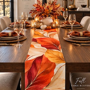 May include: A rectangular table runner featuring a printed pattern of large autumn leaves in shades of red, orange, and cream. The runner measures 40.64 cm by 228.6 cm and is displayed on a wooden dining table set with plates, glasses, and napkins.