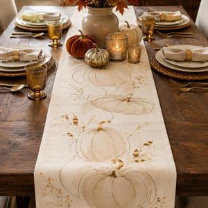 May include: A long, off-white table runner featuring a repeating print of line-drawn pumpkins and delicate botanical sprigs in a soft brown tone. The runner is centred on a dark wooden dining table set with plates, napkins, and glassware.