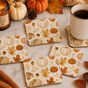 May include: Four square coasters featuring a repeating pattern of white and orange pumpkins, brown acorns, and autumn leaves in shades of gold and rust. The coasters have a light-coloured base and are arranged on a surface with a candle and a mug.