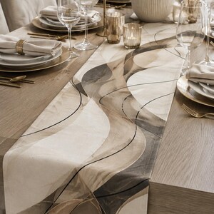 May include: A rectangular table runner featuring an abstract pattern of overlapping curved shapes in shades of beige, cream, taupe, and dark brown, accented by thin black lines that create a flowing, layered visual effect across the fabric surface.