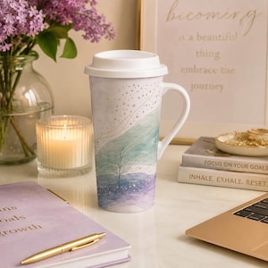 May include: A tall white ceramic travel mug with a handle and a white silicone lid. The mug features a watercolor-style design in shades of blue, green, and purple, accented with delicate gold line art of a small plant and scattered dots.