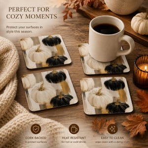 May include: A set of four square coasters featuring a printed design of white and black pumpkins with gold brushstroke accents. Each coaster has a smooth surface and a cork backing. The coasters are arranged on a wooden surface near a ceramic mug.
