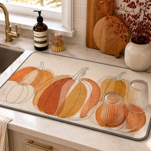 May include: A rectangular kitchen drying mat featuring a beige background with a repeating line art and watercolor-style illustration of pumpkins in shades of orange, yellow, and cream. The mat has a dark gray border and two clear ribbed glasses.