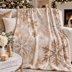 May include: A plush throw blanket featuring a cream-coloured base with a repeating pattern of large, intricate snowflakes in metallic gold and shimmering silver tones, accented with small gold dots scattered across the fabric surface.