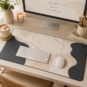 May include: A rectangular desk mat featuring an abstract design with flowing black and beige organic shapes. The surface has a smooth texture with rounded corners, providing a workspace for a white wireless keyboard and a white computer mouse.