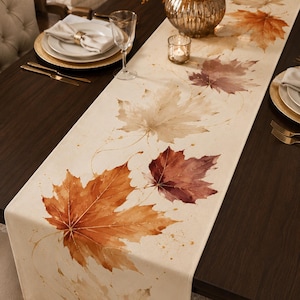 May include: A long, rectangular table runner in an off-white fabric featuring a printed watercolor design of scattered autumn maple leaves in shades of burnt orange, deep red, and muted beige, extending along the length of the textile.