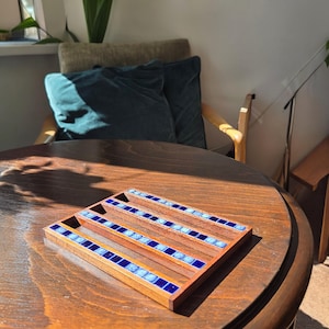 May include: A wooden tray with a rectangular frame and four parallel grooves. Each groove is lined with small, square blue tiles. The tray sits on a round, dark wooden table, with a chair and pillows in the background.