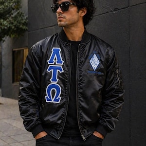 Può includere: Un giubbotto bomber in raso nero con le lettere greche Alpha Tau Omega in blu e bianco sul petto destro. Uno stemma a forma di diamante con ricami blu e bianchi si trova sul petto sinistro. La giacca presenta una chiusura con zip frontale.