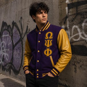 Könnte beinhalten: Eine violette Collegejacke mit goldenen Lederärmeln sowie geripptem Kragen, Bündchen und Saum mit goldenen Streifen. Auf der linken Brust sind die griechischen Buchstaben Omega, Psi und Phi in Gold gestickt, ergänzt durch goldene Druckknöpfe.