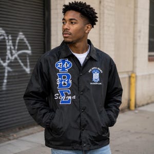 Peut inclure: Une veste coach en nylon noir portée sur un t-shirt blanc. La veste présente les lettres grecques Phi Beta Sigma en bleu avec des contours blancs, ainsi que le texte Phi Beta Sigma Fraternity, Inc. et un logo écusson sur la poitrine.