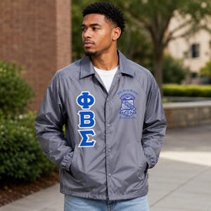 Puede incluir: Una chaqueta cortavientos gris con cuello blanco y cierre de botones a presión. El frente muestra las letras griegas ΦΒΣ en azul y blanco verticalmente en el lado derecho y un escudo azul de Phi Beta Sigma en el pecho izquierdo.