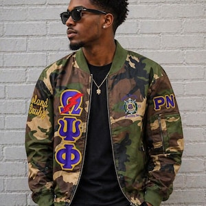 May include: A camouflage bomber jacket featuring purple and gold Greek letters on the right chest, a red lightning bolt emblem, the text Island Bruh on the right sleeve, and the letters PN on the left sleeve with a circular crest on the left chest.