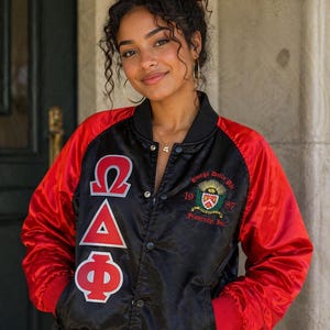 Könnte beinhalten: Eine schwarze und rote Satin-Collegejacke mit den griechischen Buchstaben Omega Delta Phi in Weiß und Rot auf der rechten Seite und einem gestickten Omega Delta Phi Fraternity, Inc. Wappen mit der Jahreszahl 1987 auf der linken Seite.