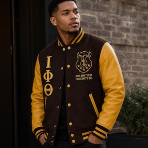 Könnte beinhalten: Eine braune Collegejacke mit gelben Lederärmeln, die auf der rechten Brust vertikal die griechischen Buchstaben Iota Phi Theta und auf der linken Brust ein kreisförmiges Wappen mit dem Text IOTA PHI THETA FRATERNITY, INC. sowie gelb gestreifte Rippbündchen aufweist.