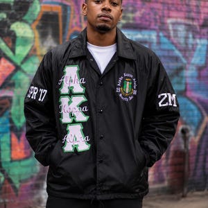 May include: A black nylon coach jacket featuring white and green embroidered lettering that reads Alpha Kappa Alpha. The jacket includes a circular crest on the chest, the text SPR '17 on the right sleeve, and the letters ZM on the left sleeve.