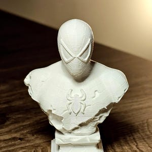 Op de afbeelding: Een witte buste van Spider-Man, met het masker en het borstembleem van het personage. De sculptuur heeft een getextureerd oppervlak en staat op een rechthoekige basis. Een verzamelobject.