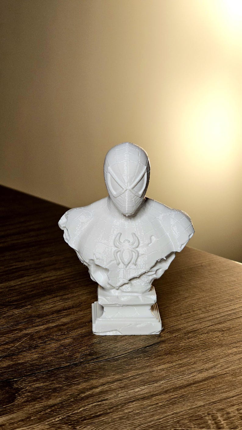 Op de afbeelding: Een witte buste van Spider-Man, met het masker en het borstembleem. De sculptuur is 3D-geprint en staat op een rechthoekige basis. De buste is een decoratief item.