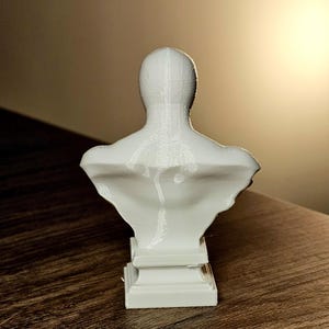 Op de afbeelding: Een witte borstbeeldsculptuur die het bovenlichaam van een persoon afbeeldt. De sculptuur is gemonteerd op een rechthoekige basis en is gemaakt van een glad, stevig materiaal. De buste staat tegen een neutrale achtergrond.