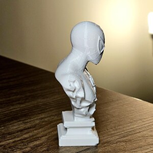 Op de afbeelding: Een witte 3D-geprinte buste van Spider-Man, met het masker en bovenlichaam van het personage. De sculptuur is gemonteerd op een rechthoekige basis en staat op een houten oppervlak.