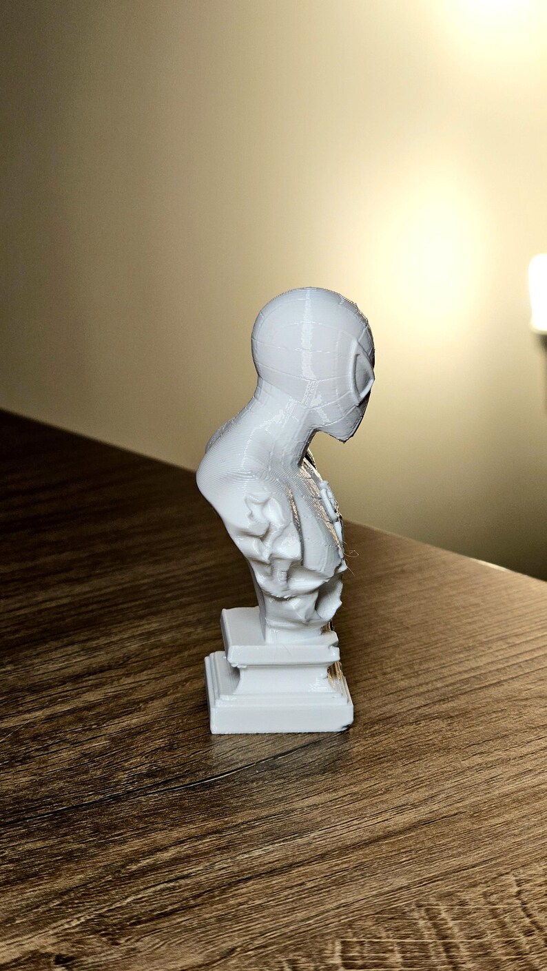Op de afbeelding: Een witte 3D-geprinte buste van Spider-Man, met het masker en bovenlichaam van het personage. De sculptuur is gemonteerd op een rechthoekige basis en staat op een houten oppervlak.