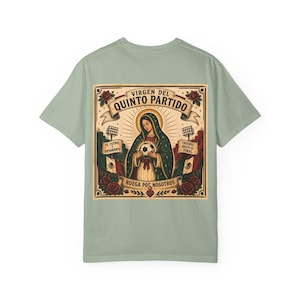 May include: A light green t-shirt featuring a detailed graphic on the back. The graphic includes an illustration of a religious figure holding a soccer ball, surrounded by text and decorative elements. The text includes the words "Virgen del Quinto Partido" and "Ruega Por Nosotros".