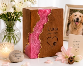 Puede incluir: Una urna rectangular de madera para mascotas con una incrustación vertical de resina rosa ondulada y bordes dorados. La parte frontal está grabada con el nombre Luna, los años 2008-2023 y un corazón con una huella de pata.