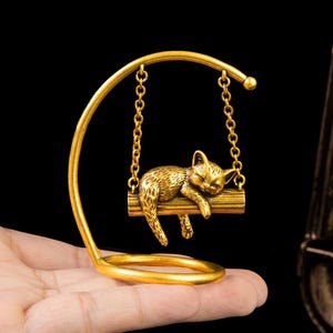 May include: A gold-toned metal ornament featuring a small cat figurine resting on a horizontal bar suspended by two chains from a curved stand. The cat is depicted in a sleeping position with its body draped over the bar, showcasing a textured finish.