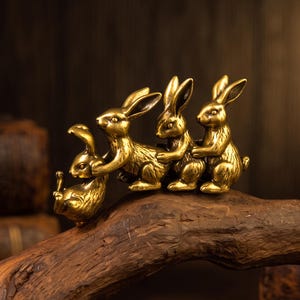 May include: A metallic gold-toned brass figurine featuring four small rabbits linked together in a line. The rabbits are depicted in a seated or crouching position, with each one holding onto the rabbit in front of it, creating a continuous chain.