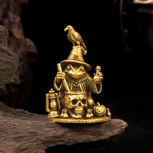 Puede incluir: Una figura de latón dorado que representa a una rana vestida de mago, con un sombrero puntiagudo y un pájaro posado encima. La rana sostiene una varilla sobre un caldero con forma de calavera, junto a una linterna y una pequeña calabaza.