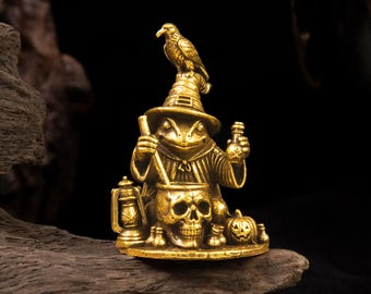 Puede incluir: Una figura de latón dorado que representa a una rana vestida de mago, con un sombrero puntiagudo y un pájaro posado encima. La rana sostiene una varilla sobre un caldero con forma de calavera, junto a una linterna y una pequeña calabaza.