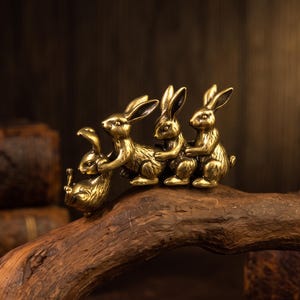 May include: A metallic brass-toned figurine featuring four small rabbits linked together in a line. The rabbits are depicted in a seated or crouching posture, with each one holding onto the rabbit in front of it, creating a continuous chain.