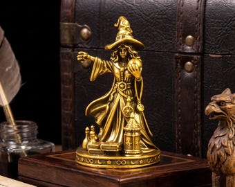 Puede incluir: Una figura de color latón que representa a un personaje con túnica y sombrero puntiagudo, sosteniendo una esfera en una mano y gesticulando con la otra. La base presenta estrellas, lunas y símbolos de infinito grabados, con libros y una linterna.