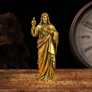 Könnte beinhalten: Eine goldfarbene Metallfigur von Jesus Christus, die in einem drapierten Gewand steht. Die Figur hat langes Haar, einen Bart und ein Herz-Jesu-Emblem auf der Brust. Die rechte Hand ist erhoben, wobei zwei Finger zum Segen ausgestreckt sind.
