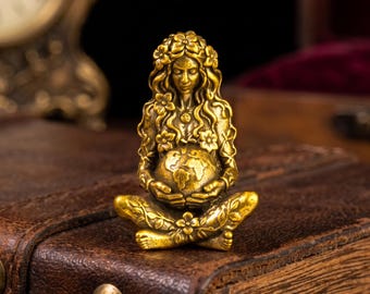 Puede incluir: Una pequeña figura de latón en tono dorado que representa a un personaje sentado con el pelo largo y fluido adornado con flores. La figura está en posición de piernas cruzadas, sosteniendo un globo esférico con motivos florales grabados.