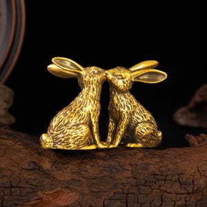 May include: A pair of metallic gold-toned rabbit figurines positioned facing each other. The rabbits feature detailed, textured fur patterns, long upright ears, and are depicted in a seated posture. The sculpture has a polished finish.