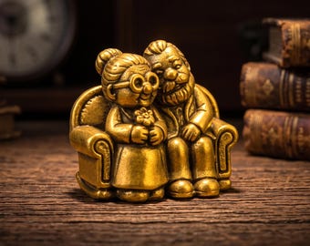 Puede incluir: Una figura de metal en tono dorado que representa a una pareja de ancianos sentados juntos en un sofá pequeño. La figura de la izquierda lleva gafas redondas y sostiene una flor, mientras que la de la derecha tiene barba y apoya un brazo en el respaldo.