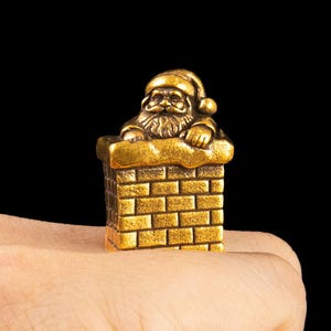 Könnte beinhalten: Ein goldfarbener Metallring mit einer dreidimensionalen Figur des Weihnachtsmanns, der aus einem Backsteinkamin hervorschaut. Die Weihnachtsmannfigur hat einen detaillierten Bart und eine Mütze, während der Kamin ein Ziegelmuster aufweist.