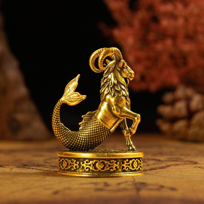 May include: A metallic gold-colored figurine depicting a mythical creature with the upper body of a goat and the lower body of a fish. The sculpture features detailed fur, curved horns, and scaled tail textures, mounted on a circular base with ornate patterns.