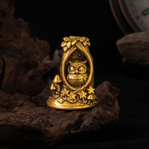 Può includere: Una piccola statuetta in metallo color oro a forma di tronco d'albero cavo. All'interno dell'apertura si trova un gufo dettagliato con grandi occhi. La base dell'albero è decorata con funghi scolpiti e foglie a forma di stella in rilievo.