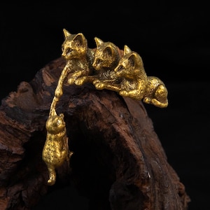 May include: A gold-toned metal figurine featuring four cats. Three cats are positioned in a row, while a fourth cat hangs vertically below, reaching up to touch the paw of one of the cats above. The metal has a textured, antique finish.