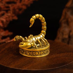 May include: A small, metallic gold-colored scorpion figurine perched atop a circular base. The base features an embossed Scorpio zodiac symbol and intricate decorative scrollwork patterns around its side, with a polished finish.